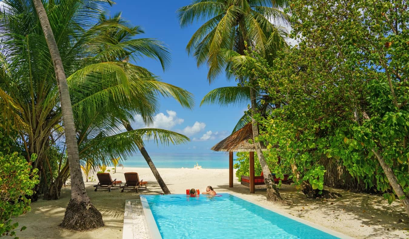 People relaxing in a long rectangular pool surrounded by palm trees and the ocean in the distance in the Maldives