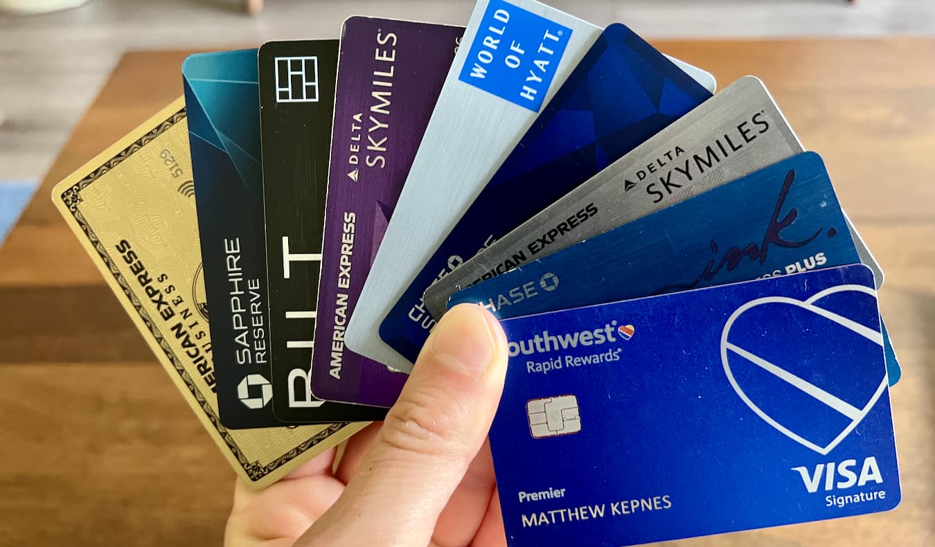 A hand holding a fan of 9 different US travel credit cards, including American Express Gold, BILT, Chase Sapphire Reserve, World of Hyatt, Delta SkyMiles, Southwest Rapid Rewards