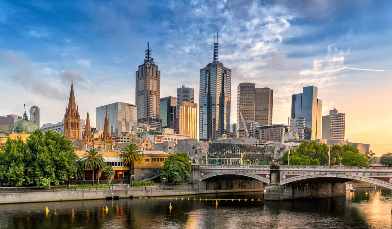 The towering skyline of Melbourne, Australia with trees and a bridge in the foreground near the river