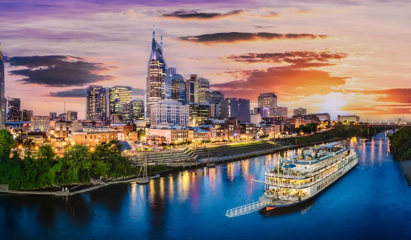 The skyline of Nashville, TN at dusk with an old boat cruising the river nearby