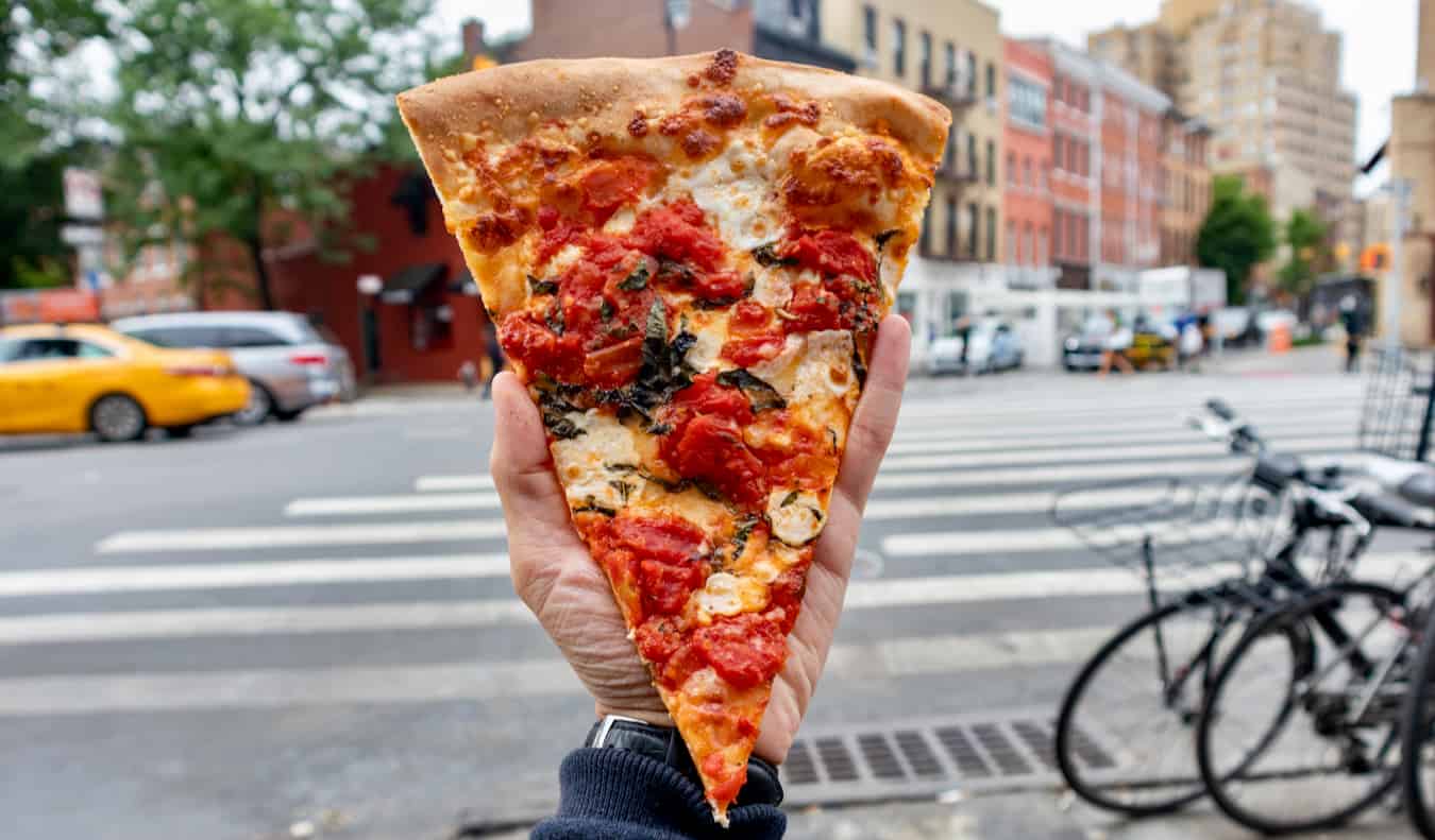 A hand holding up a slice of pizza on a NYC street