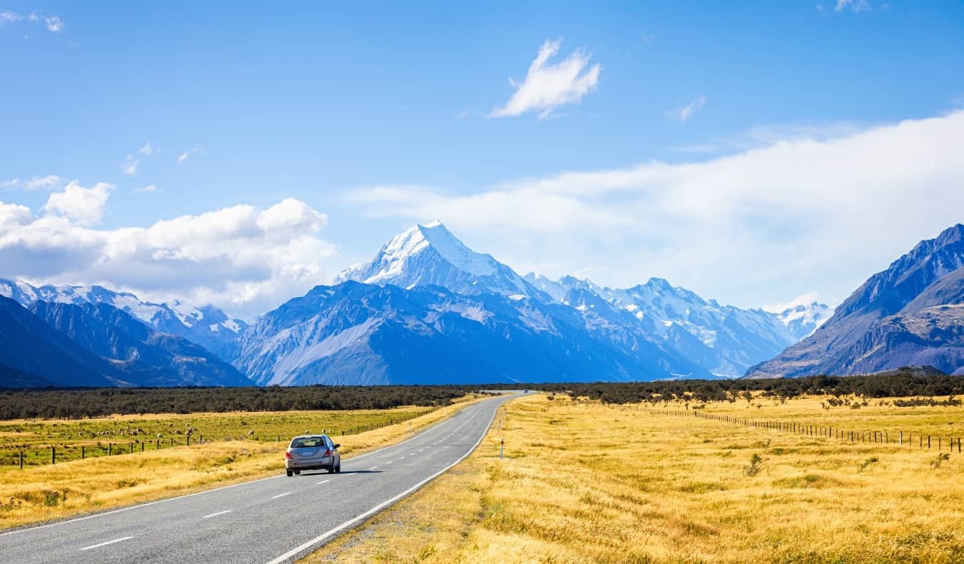 Car driving down a road with golden fields on either side, leading towards snow-capped mountains on the South Island of New Zealand