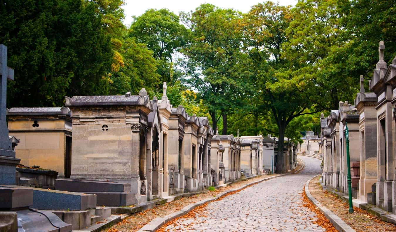 Mausoleums lined up along a cobblestone pathway littered with orange leaves at Pere Lachaise Cemetery in Paris, France