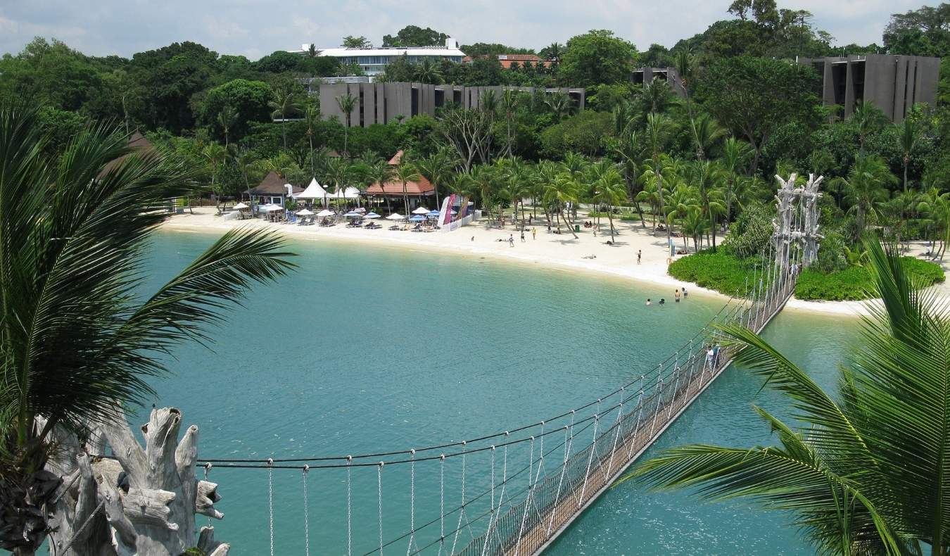 A hanging pedestrian suspension bridge leading to a sandy beach lined with palm trees and small huts in Sentosa, Singapore