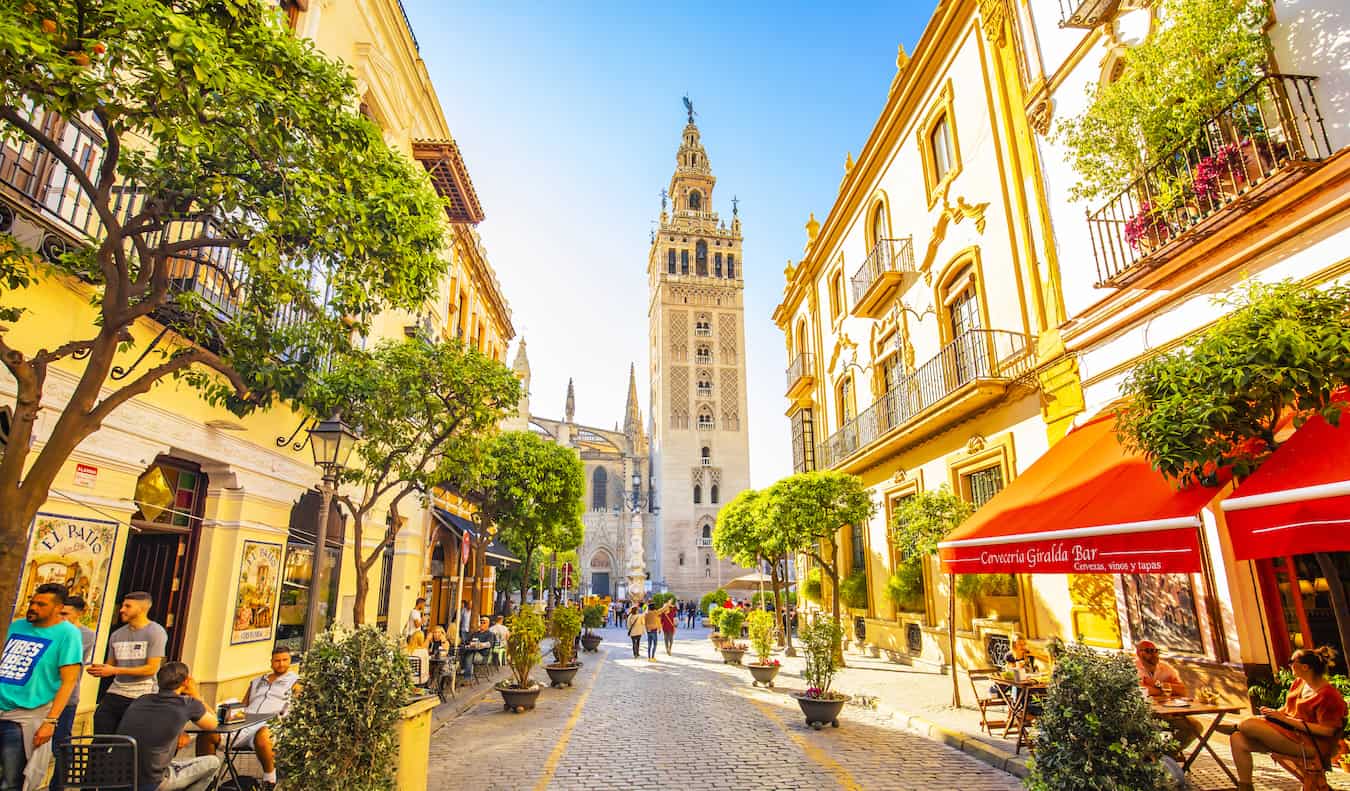 People walking along a quiet, narrow street in sunny Sevilla, Spain with a church tower in the distance