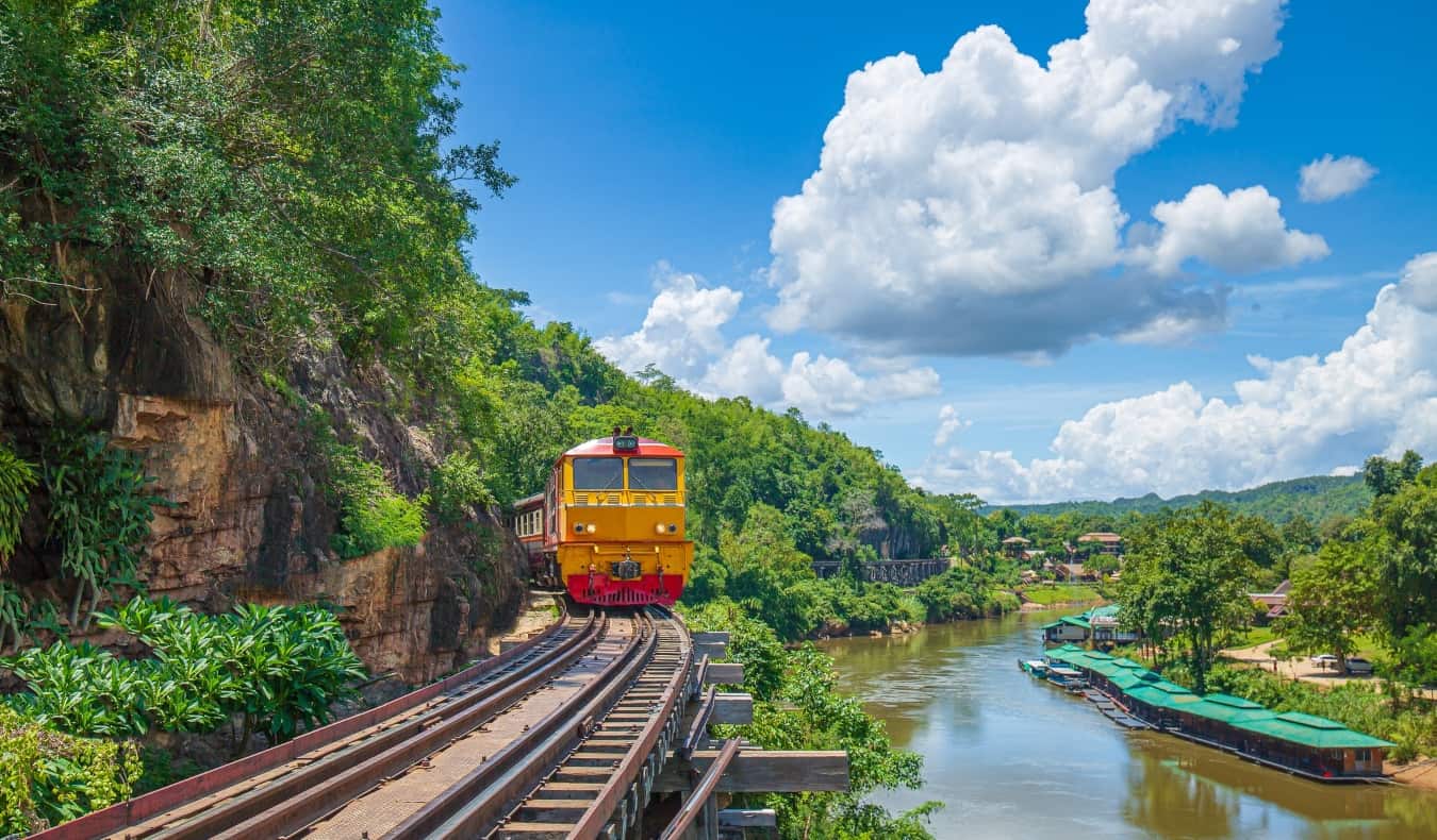 a train hugging the cliff side in Kanchanaburi, Thailand with on a sunny day with beautiful views over Kwai Noi River on the right
