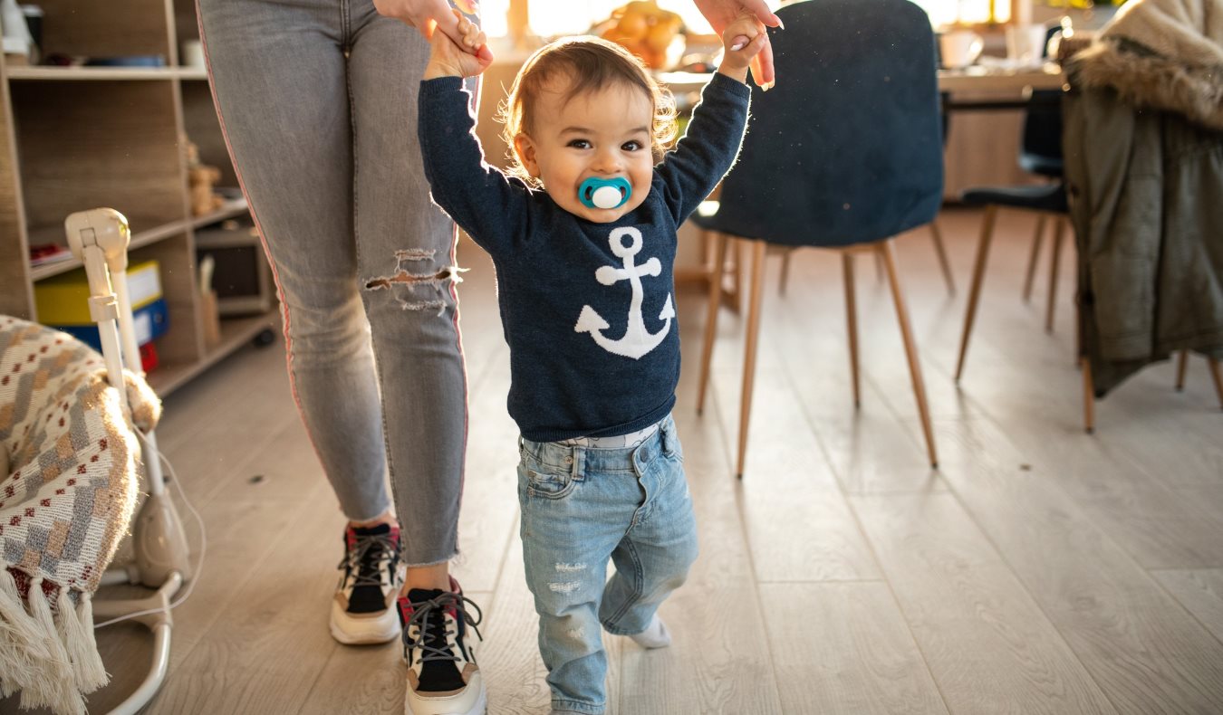 An au pair standing holding a toddler's hands as they run through a living room