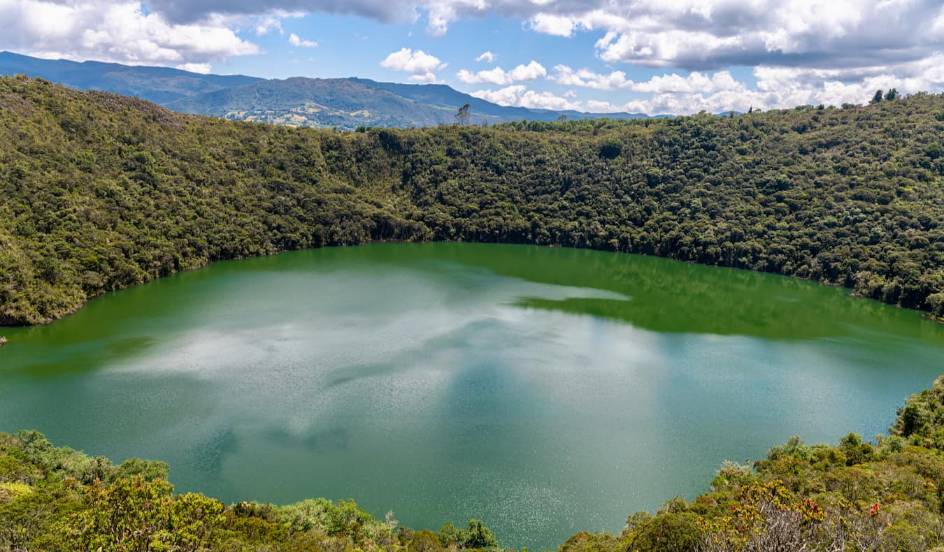 The emerald green Lake Guatavita surrounded by lush ferns, greenery, and rolling hills, in Colombia