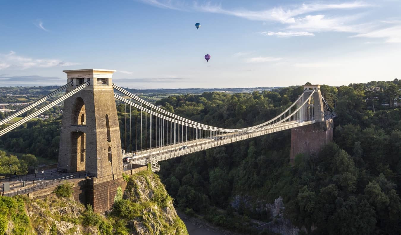 Looking out at the Clifton Suspension Bridge that cuts over a deep gorge, with a few hot air balloons in the background in Bristol, UK