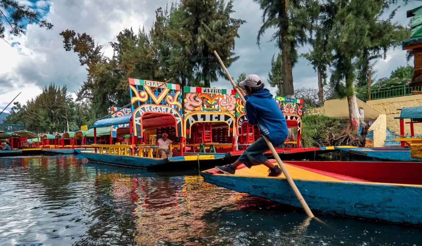 A young boy pushes a colorful boat down a river with a long stick in the Xochimilco Canals in Mexico City, Mexico