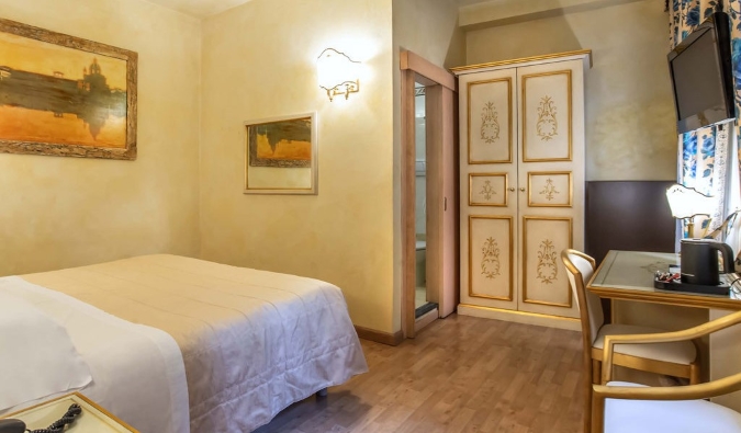 A hotel room with a simple double bed, a desk, paintings of Florence on the soft yellow walls, and a gilded wardrobe in the corner at Hotel Alba Palace in Florence, Italy