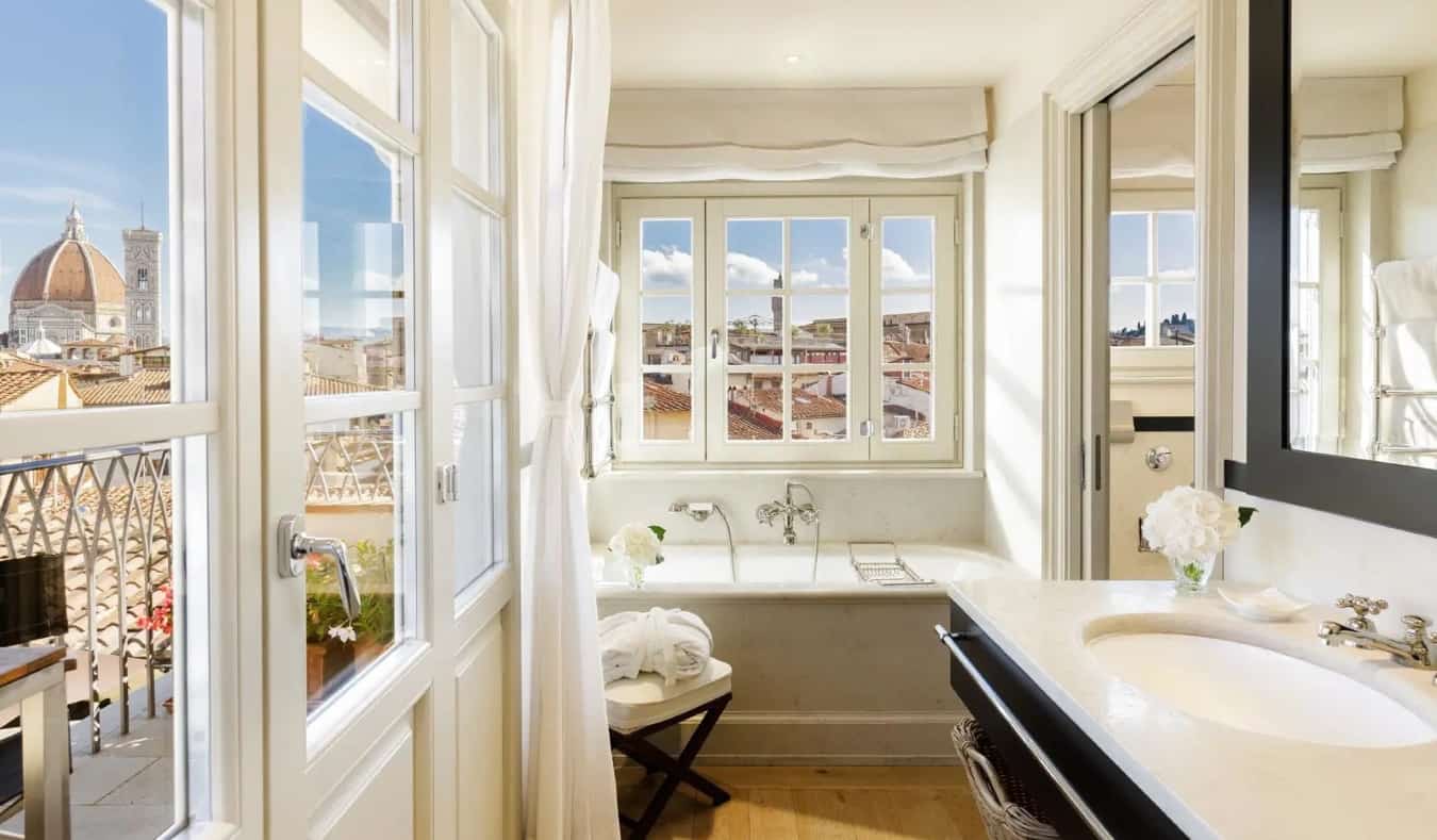 A luxurious bathroom filled with a large bathtub, wooden floors, and windows with a view of the Duomo out of one of them at The Place, a five-star hotel in Florence, Italy