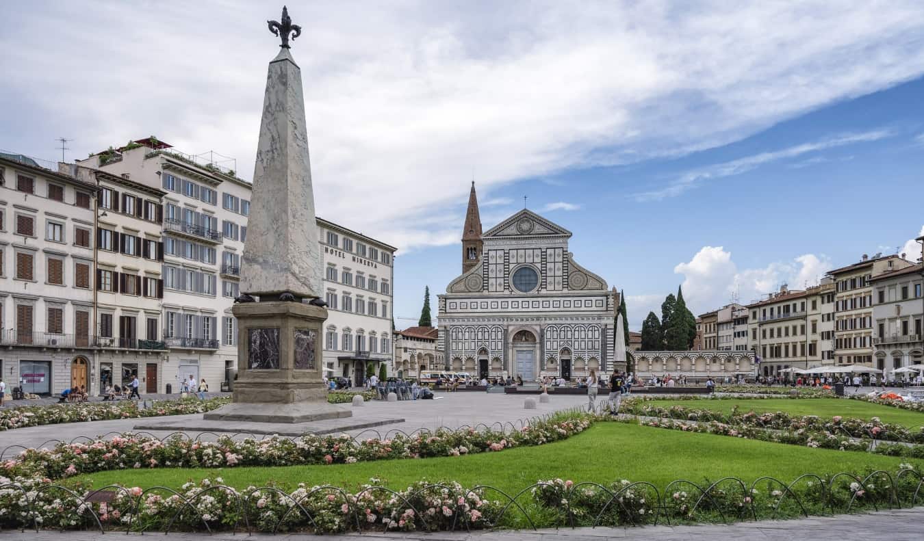 A piazza with flowers and grass around a monument and the painted Santa Maria Novella basilica in the background in Florence, Italy