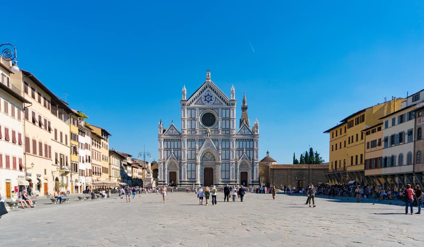 Expansive piazza lined with buildings, with people walking around in the open space in front of the majestic painted Santa Croce Basilica in Florence, Italy