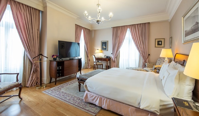 Um grande quarto de hóspedes com janelas adornadas com cortinas rosa, pisos de madeira e outros elementos históricos no Pera Palace Hotel em Istambul, Turquia