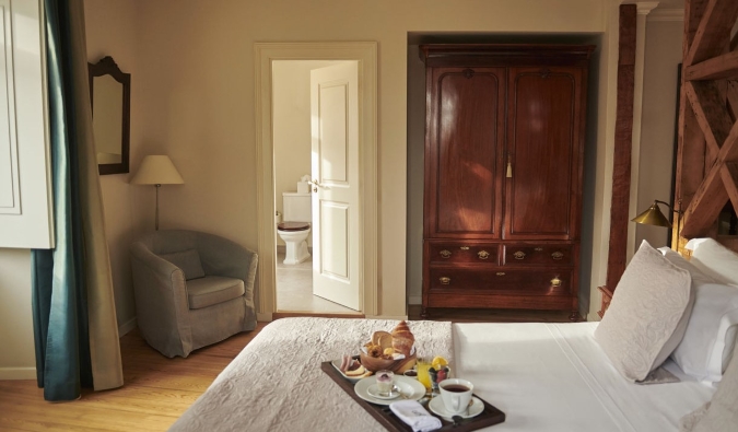 A guest room with a large wooden wardrobe, hardwood floors, a bed with a tray of breakfast foods on it, and a small armchair in the corner at Hotel Alegria in Lisbon, Portugal