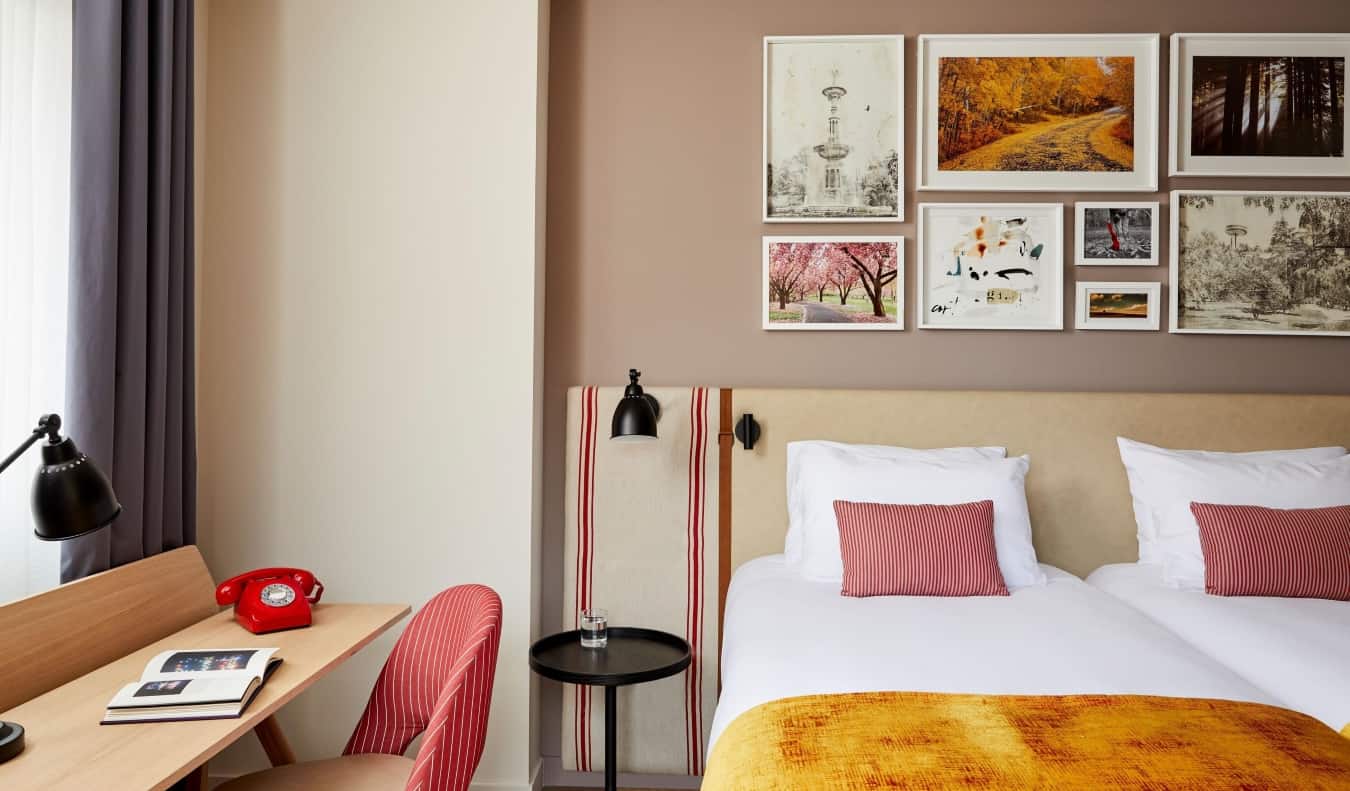 A cluster of photographs and drawings hang above a bed with red pillows and orange blankets, next to a wooden desk with a red rotary phone on it at Hotel Indigo Madrid-Princesa in Madrid, Spain