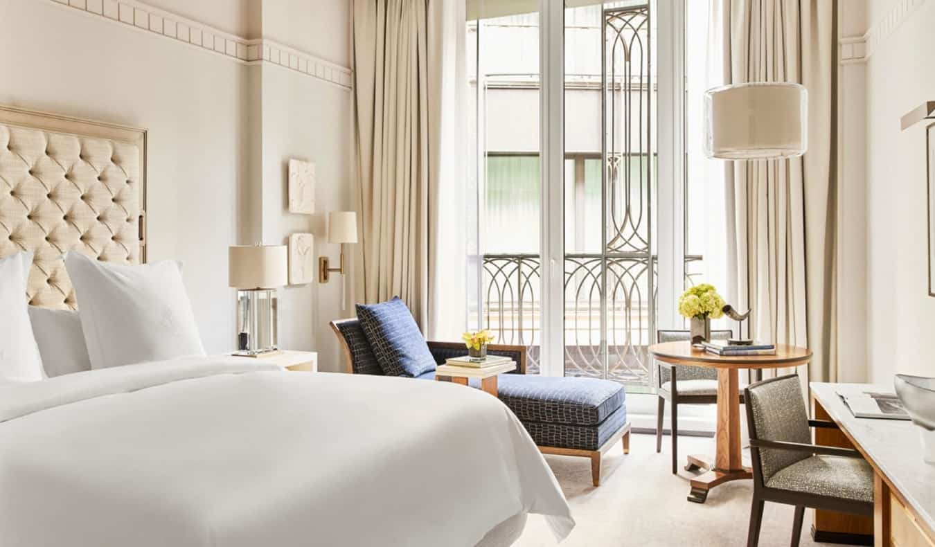 A luxurious cream-colored hotel room with a large bed, blue chaise longue by the open window, and a desk at the Four Seasons in Madrid, Spain