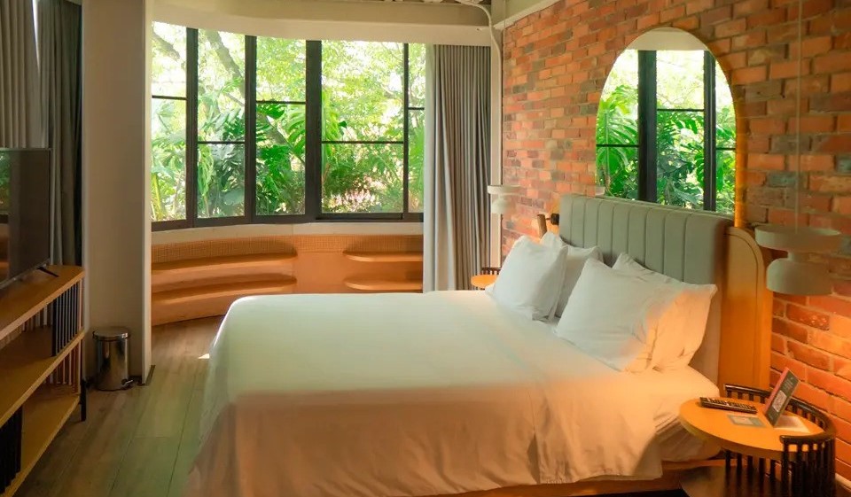 A cozy guest room with wooden floors, an exposed brick wall, and a large window opening to leafy greenery, in Medellin, Colombia