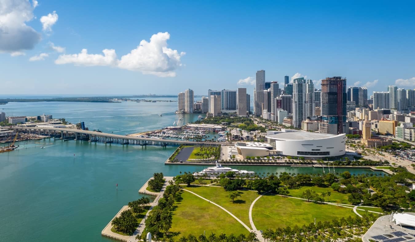 Aerial shot of Downtown Miami with its many skyscrapers, with a sprawling park in the foreground and a long bridge going to an island underneath a blue sky