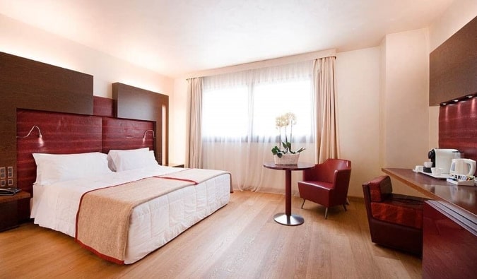 A king-sized bed in a spacious hotel room with a desk and small table with flowers, all decorated in wood and red accenting at Art Hotel Navigli, a hotel in Milan, Italy