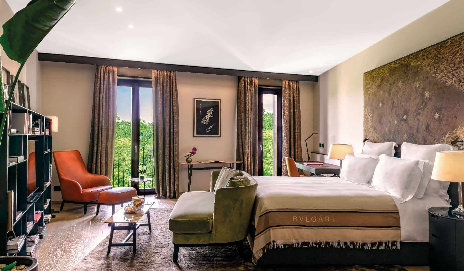 A luxurious hotel room with a velour sofa, leather armchair, bookcases, and king-sized bed covered in blankets at Bulgari Hotel Milano, a five-star hotel in Milan, Italy