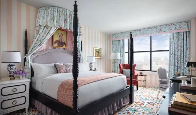 Quarto de hóspedes no Graduate Hotel em Nashville, com um retrato de Dolly Parton sobre a cabeceira de uma cama de dossel, paredes listradas de bengala de doces, um carpete estampado e grandes janelas que deixam entrar muita luz natural