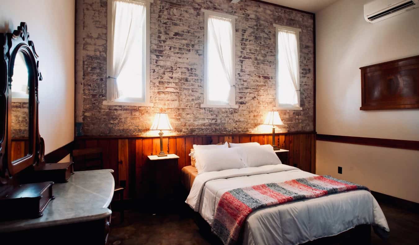 A queen bed in a guest room with exposed brick walls and antique furniture at The Inn at the Old Jail, a hotel in New Orleans, Louisiana