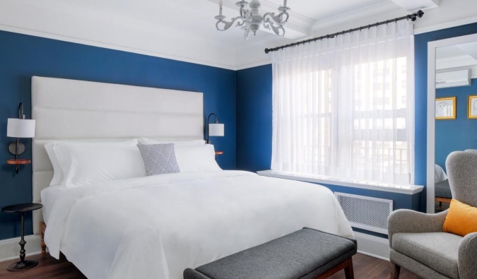 Simple hotel room with deep blue walls, a chandelier, and sitting chair next to a queen-sized bed at voco The Franklin hotel in NYC