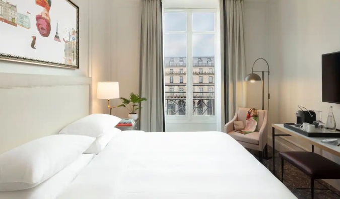 A Parisian hotel room with a queen sized bed, painting on the wall, and an open window showing the iconic architecture of Paris in the background