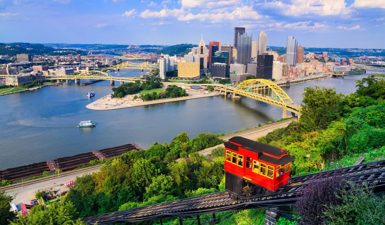 A red funicular car going up the mountain in the foreground with the skyline of Pittsburgh, PA with its many bridges spanning the river in the background