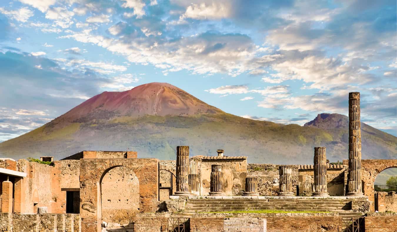 The sweeping vista overlooking Pompeii, Italy with ancient ruins in the foreground surrounded by lush grass and greenery, and Mount Vesuvius in the background