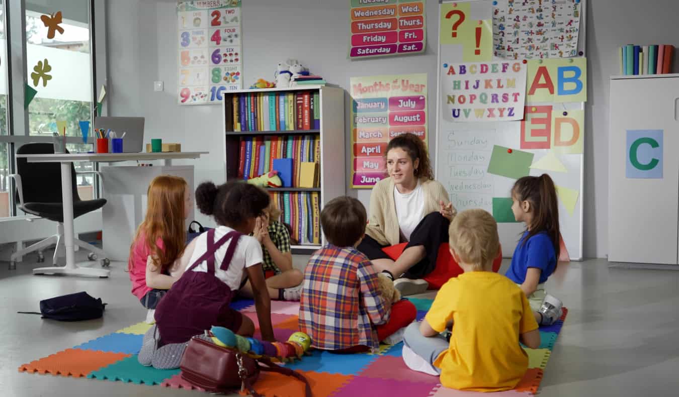 A female teacher sits on the ground with a circle of small children in front of her, with English learning materials on the boards behind her