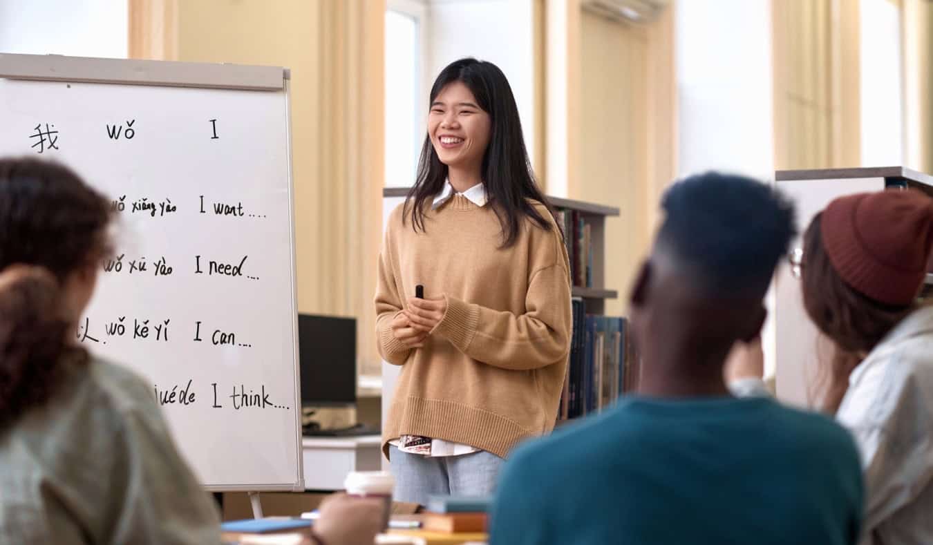 A young Asian woman in front of a white board, teaching Chinese to a group of people sitting at a table