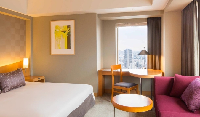 A hotel room with a double bed, a wooden desk, chair, and a red couch underneath a large window overlooking the skyline of Tokyo, Japan