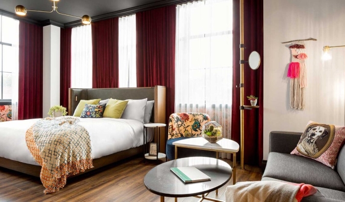 A large guestroom with a bed in front of floor-to-ceiling windows lined with rich red draperies, and a couch and table off to the side at The Broadview Hotel in Toronto, Canada
