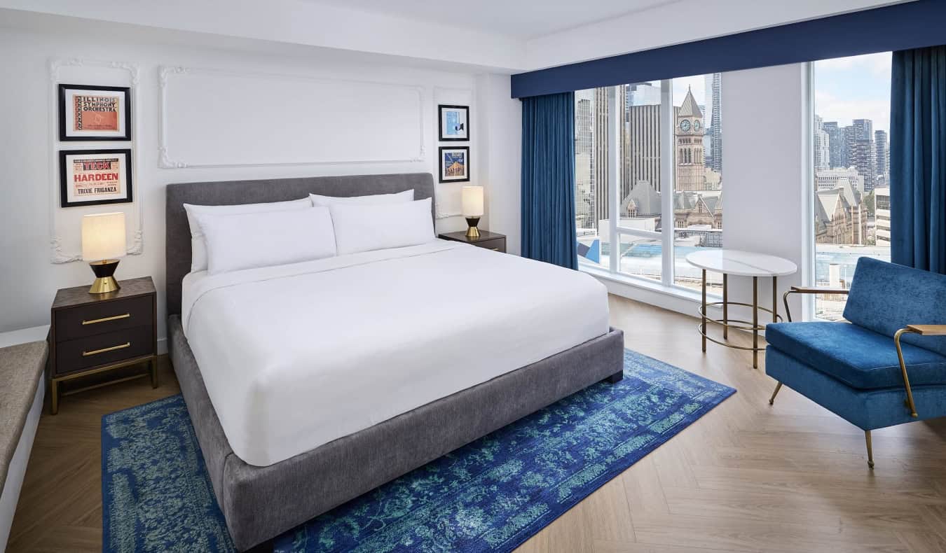 A king-sized bed with picture frames hanging from the walls on either side and large floor-to-ceiling windows overlooking the cityscape of Toronto, Canada, at the Pantages Hotel Downtown