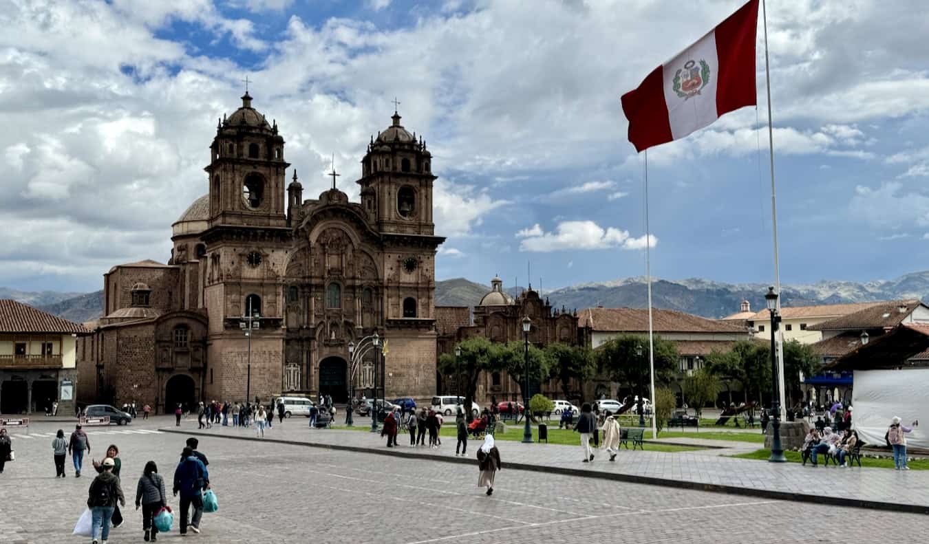 A huge flag flutters in the breeze with historical buildings nearby on a sunny day in Peru