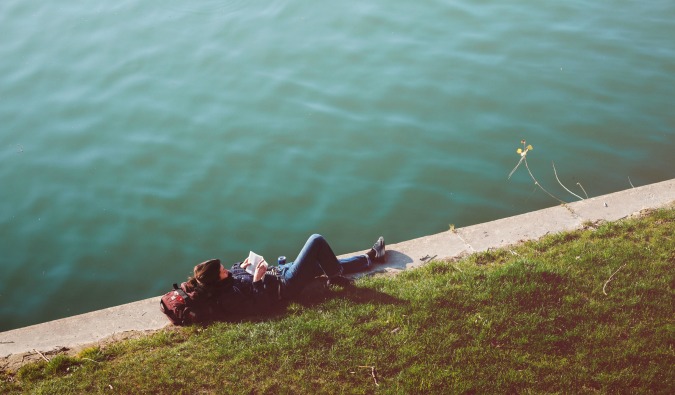 A budget traveler laying in the grass reading a book by the water