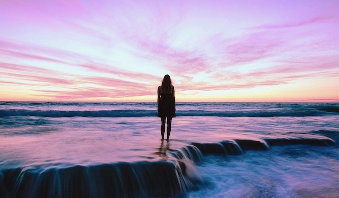 A woman watching a bright sunset while standing in shallow water on a beach