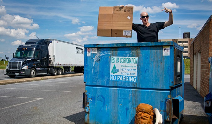 Guy in a dumpster holding up a piece of cardboard for his hitchhiking sign with a truck passing