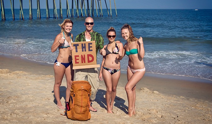 Man standing on the beach with 3 girls in bikinis and a cardboard hitchhiking sign saying THE END