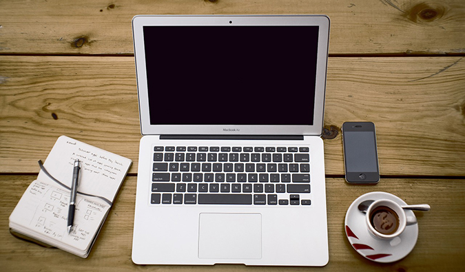 A photo of a laptop on a wooden desk with a notebook and cup of coffee