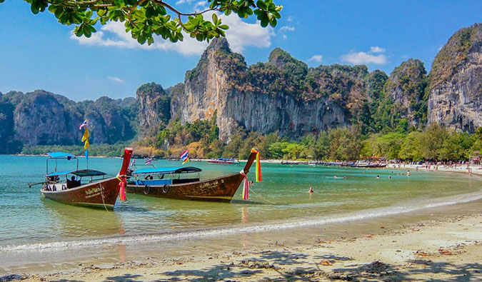 Red longtail boats pulled up to a picturesque beach with cliffs in the background, in Thailand